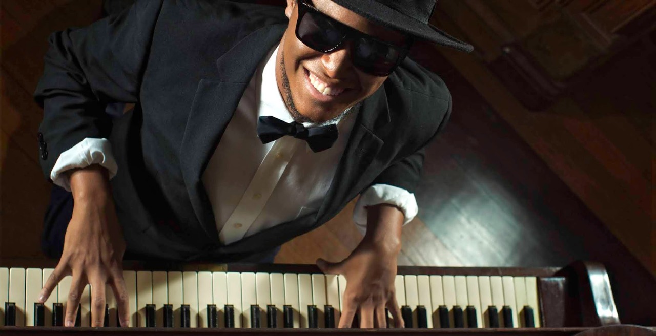 Foto do pianista Luis Otávio vestindo chapeu e terno tocando piano