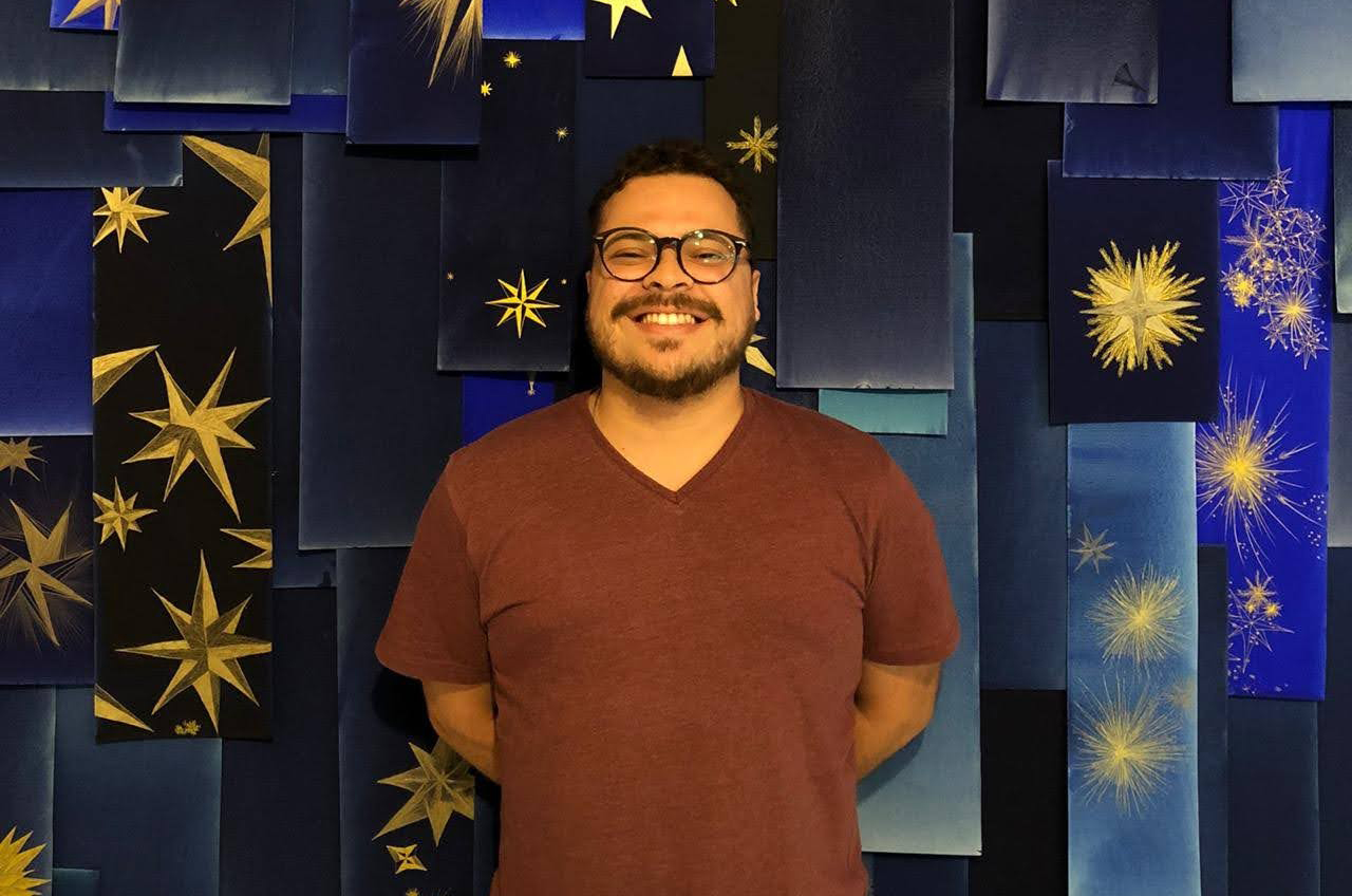 Editor do Itaú Cultural, William Nunes, em pé sorrindo com fundo azul com estrelas atrás